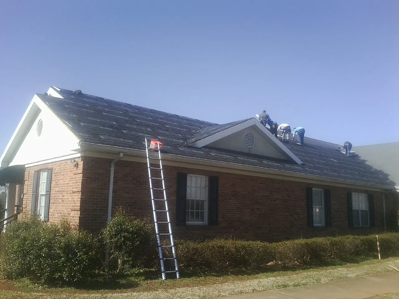 Crew tearing shingles off a brick ranch roof with a ladder set — Metal Roof Repair work by our crew in Roosevelt